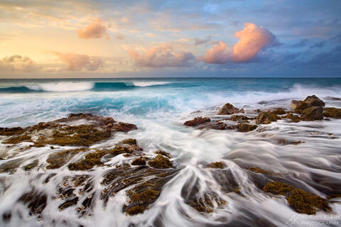 Dramatic sunrise on the south shore of Kauai island in Hawaii as water flows over rocks with a blue and yellow sky. Aqua blue water waves crash ashore. 