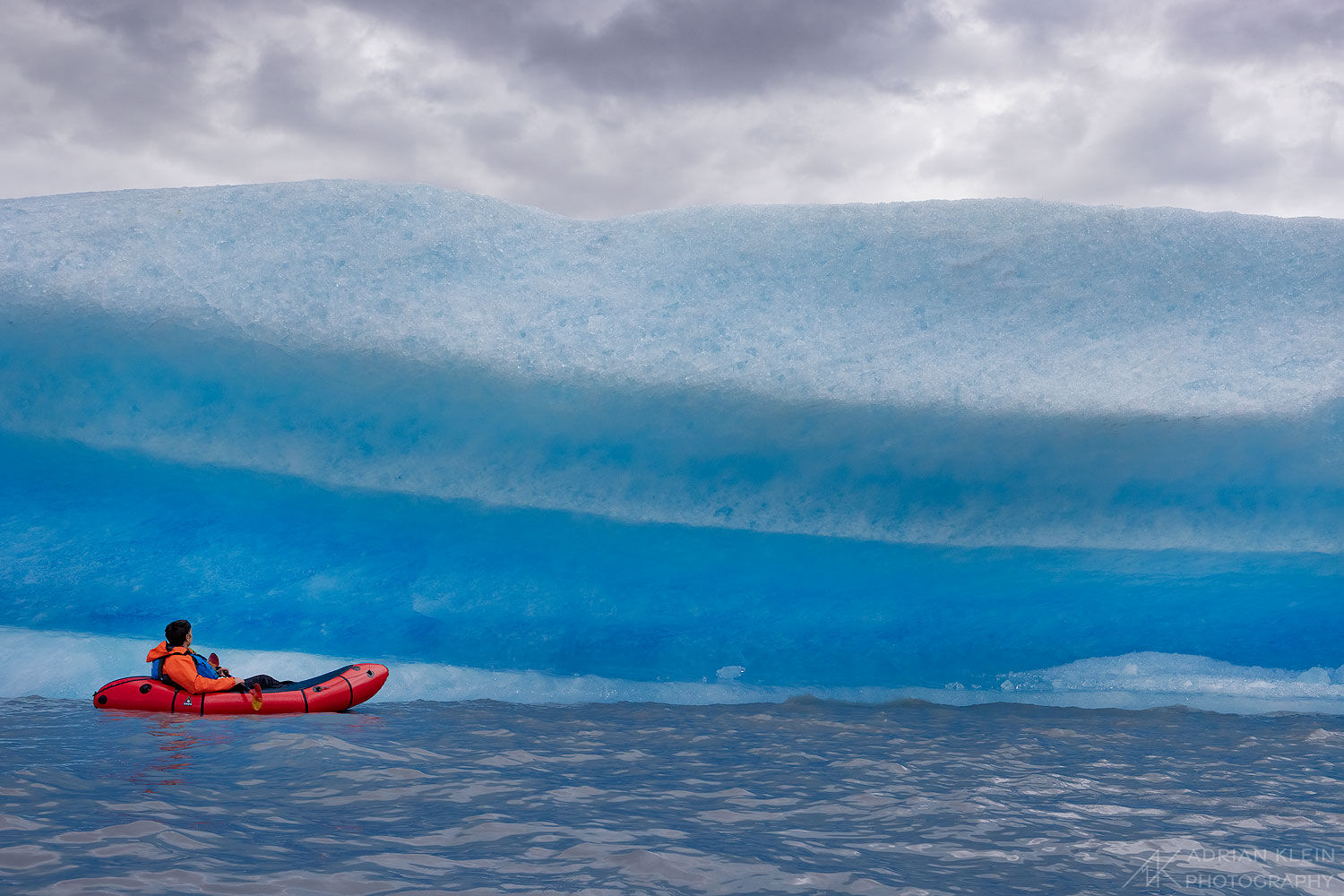 Person on pack raft floating next to large iceberg in Alaska.