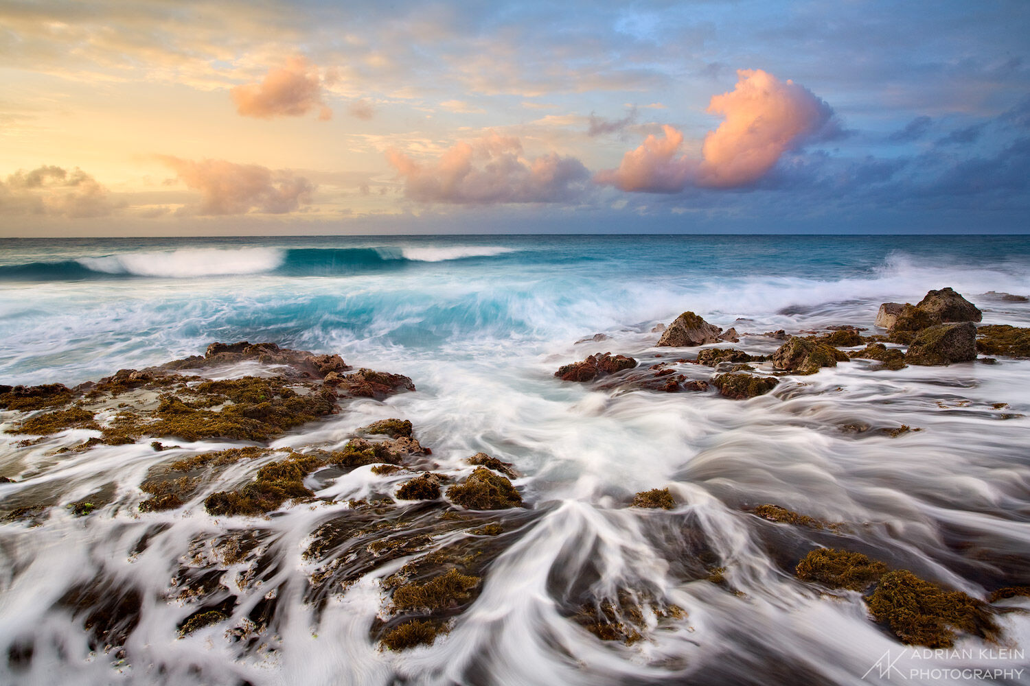 Dramatic sunrise on the south shore of Kauai island in Hawaii as water flows over rocks with a blue and yellow sky. Aqua blue water waves crash ashore.