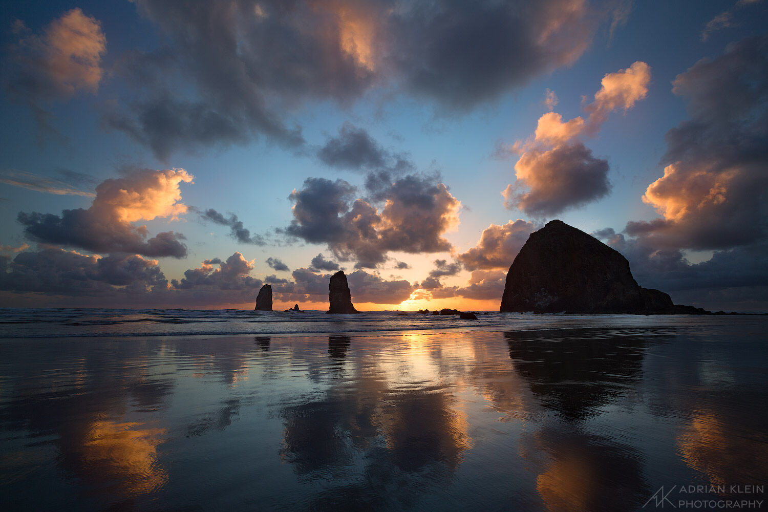 Dramatic sunset of Haystack Rock on the Oregon coast with reflections on the beach. This seascape photo feels like a dream with large puffy clouds. 